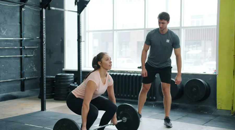 Woman deadlifting a barbell while a personal trainer coaches her in a bright Phuket gym